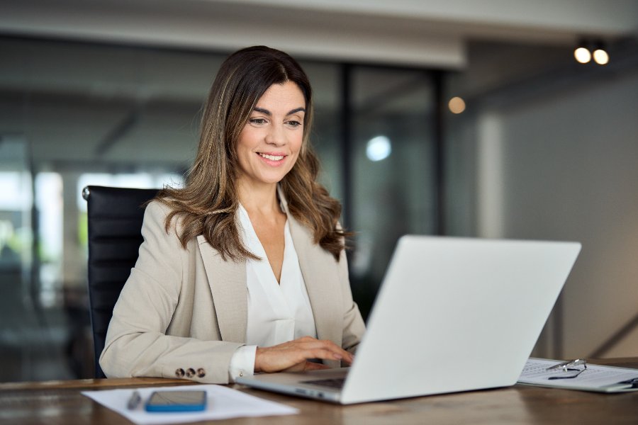 Smiling busy mature middle aged professional business woman manager executive wearing suit looking at laptop computer technology in office working on digital project sitting at desk.