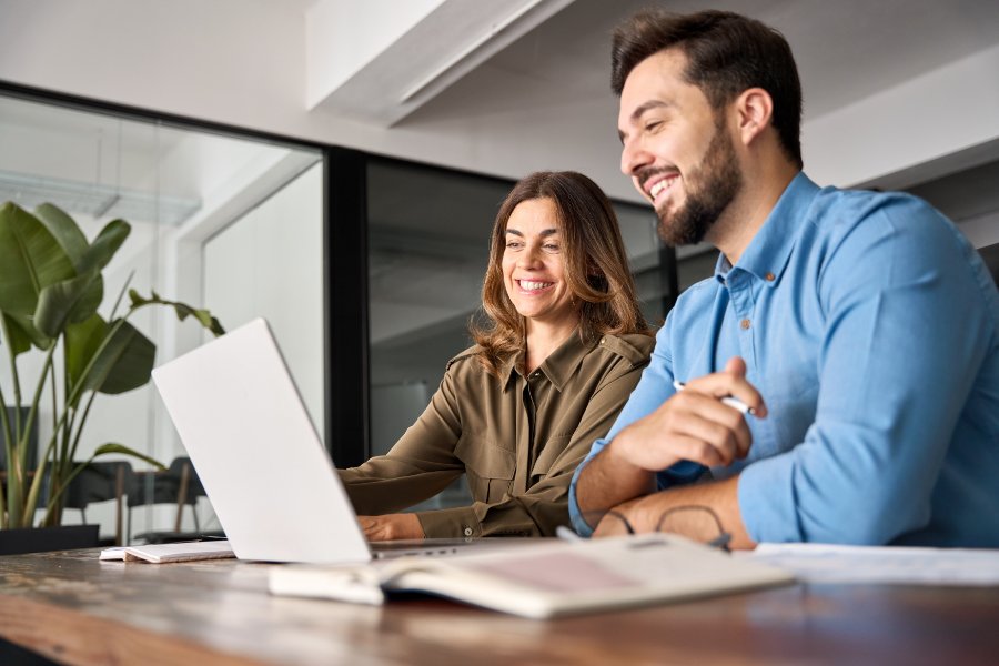 Two smiling professional business people talking using laptop computer working in office. Happy colleagues or entrepreneurs team man and woman discussing corporate technology at workplace.