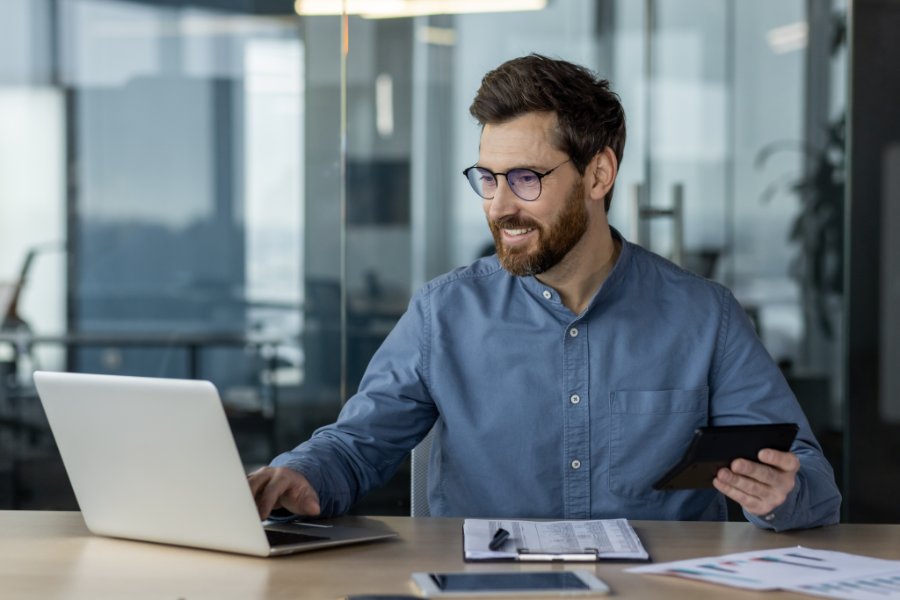 A smiling young man accountant, financial expert works in the office with documents, on a laptop, holding a calculator in his hands.
