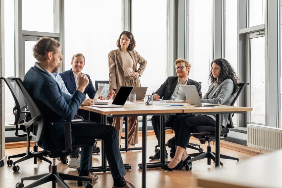 A group of business professionals are having a meeting in a modern office. The meeting is taking place around a large, rectangular table with several laptops and papers on it.